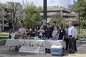 A group of people is gathered outdoors behind a table promoting Cul-de-Sac, a literary magazine at College of the Canyons. The table is covered with a banner displaying the magazine’s name and website, along with an assortment of snacks, books, and promotional materials. Some individuals in the group are wearing name tags, and one person holds up a copy of the magazine. Behind them, there are blooming trees, a modern campus building labeled "Boykin Hall," and a few other individuals in the background. The group appears cheerful, posing for a photo in a bright and lively setting.